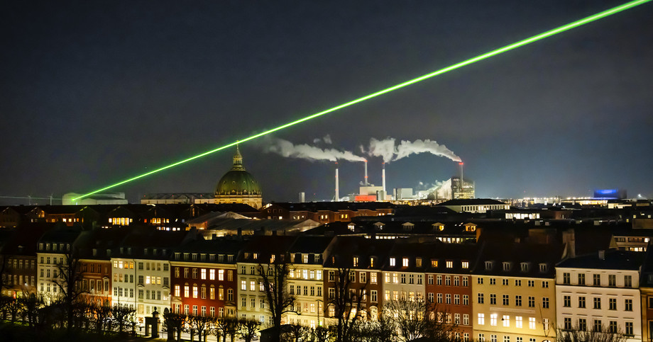 Store lysstråler skærer sig op gennem himlen og kan ses på tværs af byen og hen over byens tage. 