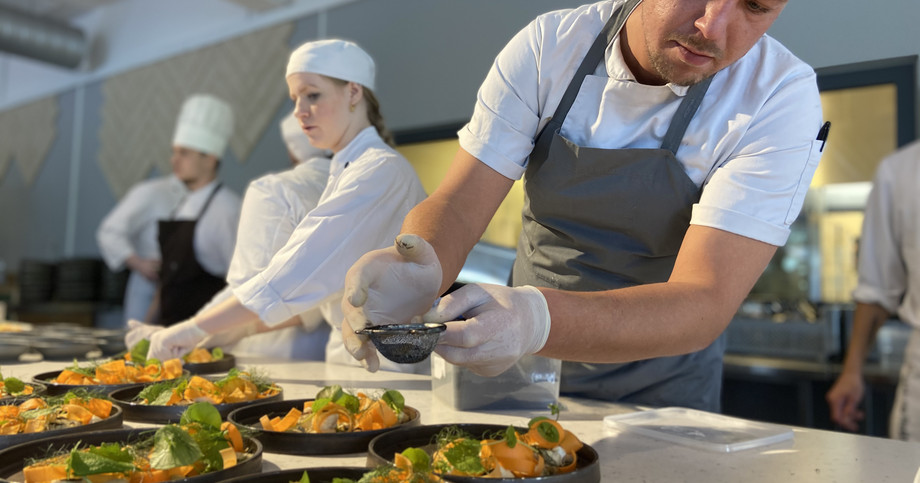 A chef plates food on a dish in a kitchen while other chefs work around him.