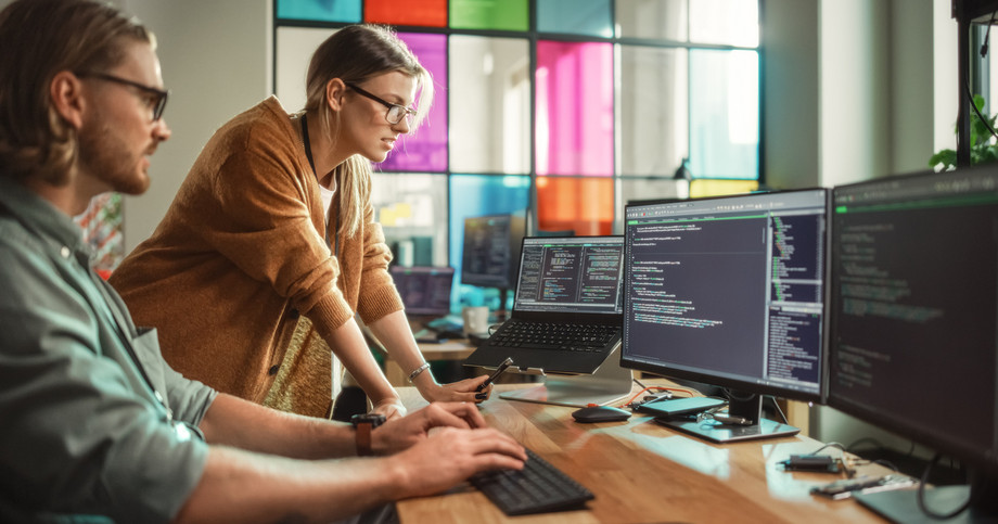 Dette billede viser to personer, der samarbejder om softwareudvikling på et kontor. En mand sidder og skriver på et tastatur, mens en kvinde står ved siden af ham, læner sig frem og peger på koden, der vises på skærmen. Begge er fokuserede på flere skærme, der viser linjer af kode. Kontoret har et moderne, åbent layout med farvede glasruder i baggrunden. Scenen formidler teamwork, problemløsning og et teknologidrevet arbejdsmiljø.