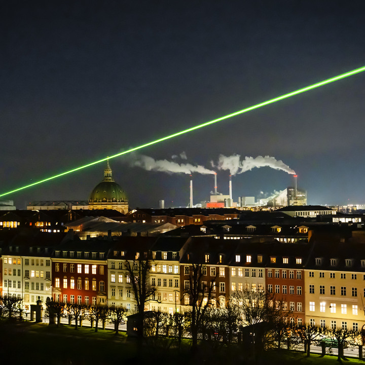 Store lysstråler skærer sig op gennem himlen og kan ses på tværs af byen og hen over byens tage. 