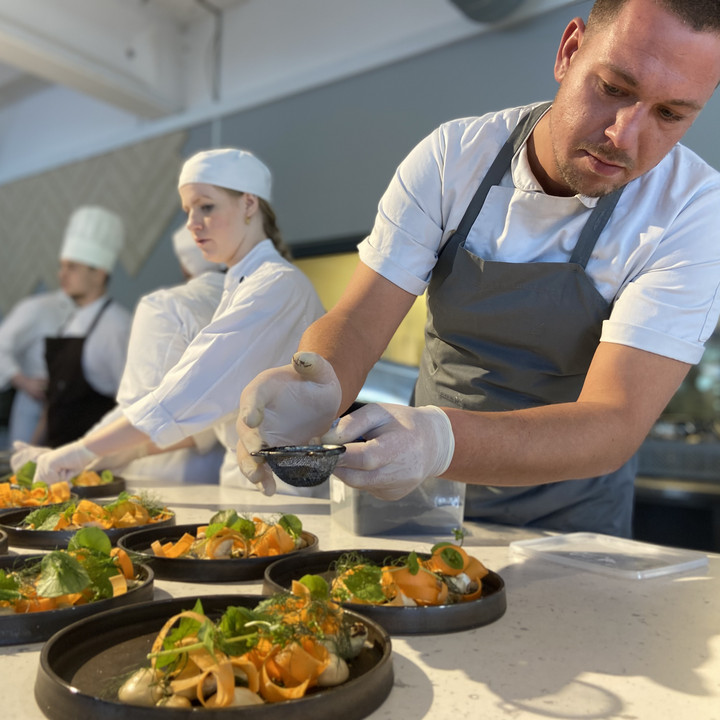 A chef plates food on a dish in a kitchen while other chefs work around him.