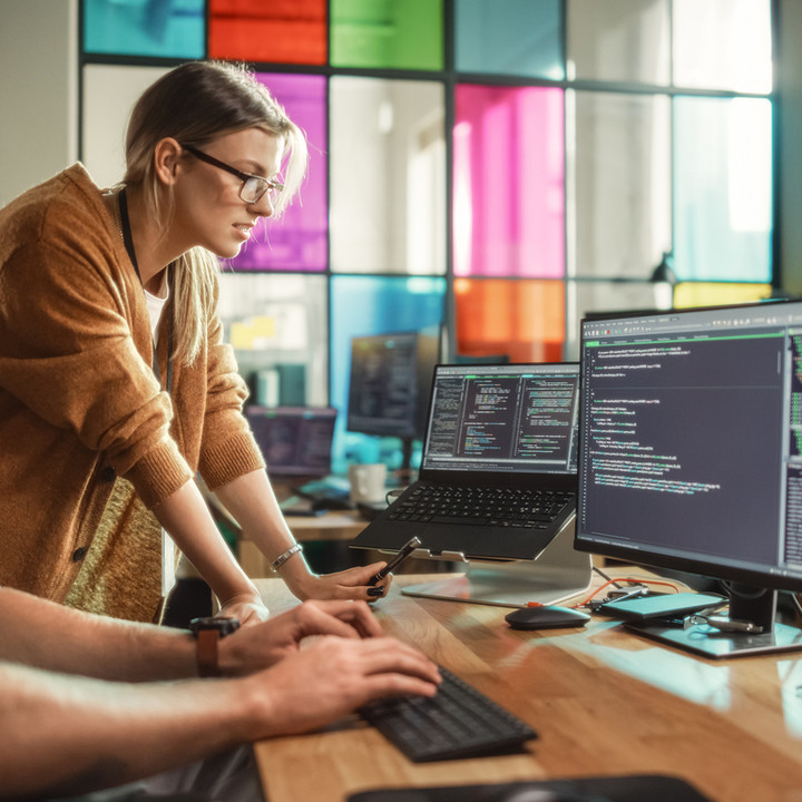 Dette billede viser to personer, der samarbejder om softwareudvikling på et kontor. En mand sidder og skriver på et tastatur, mens en kvinde står ved siden af ham, læner sig frem og peger på koden, der vises på skærmen. Begge er fokuserede på flere skærme, der viser linjer af kode. Kontoret har et moderne, åbent layout med farvede glasruder i baggrunden. Scenen formidler teamwork, problemløsning og et teknologidrevet arbejdsmiljø.